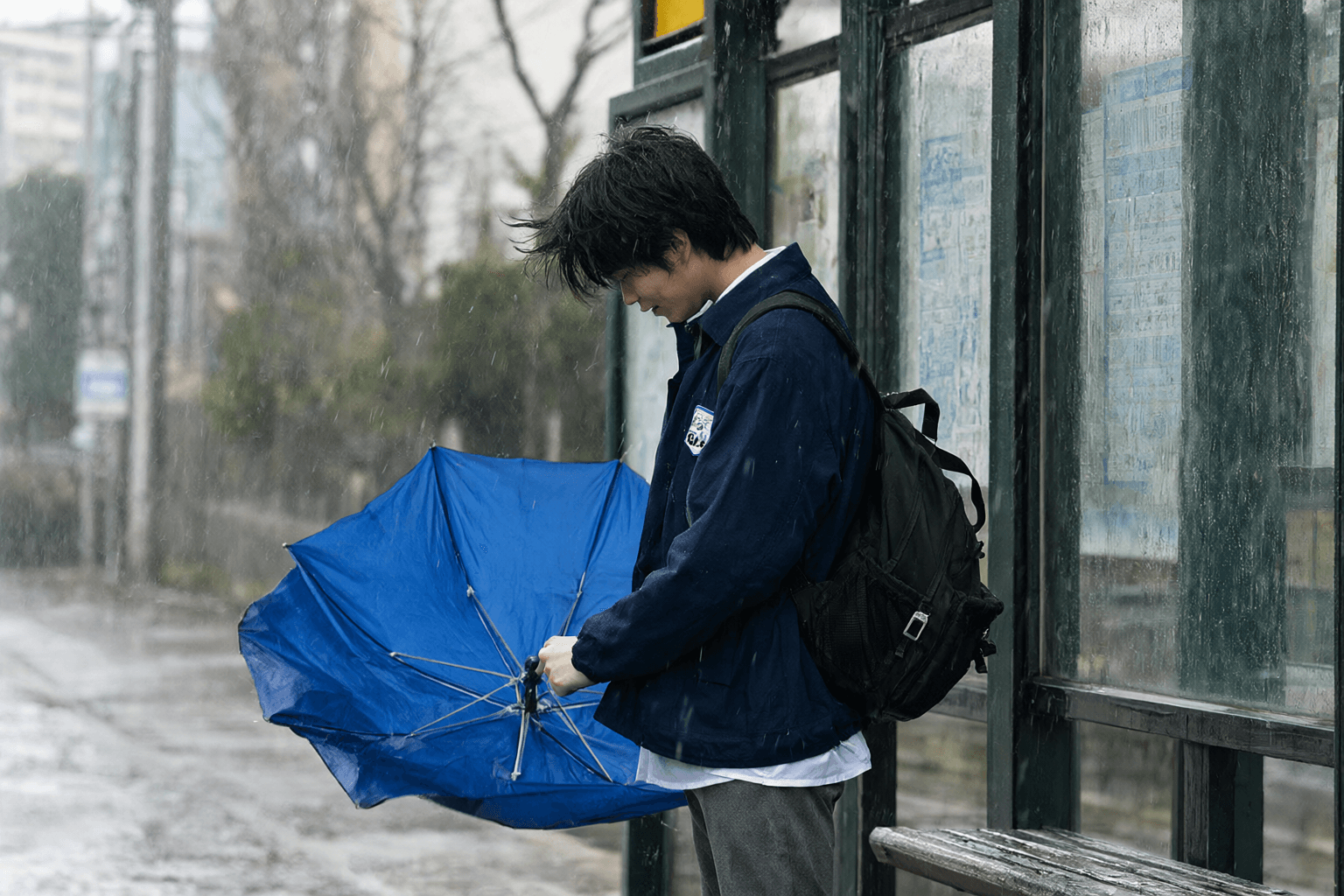 Leo is waiting at a bus stop in heavy rain. The wind is very strong and his umbrella is breaking.